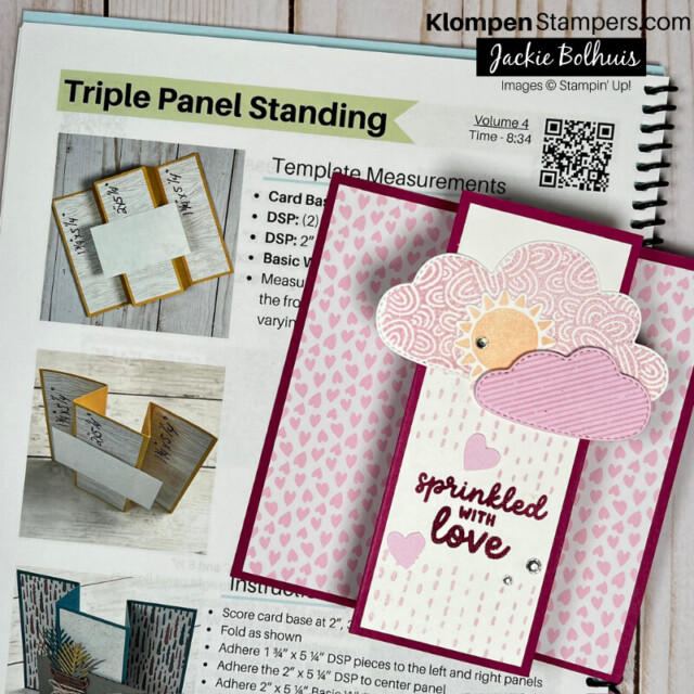 A triple panel standing fun fold card that is designed with pink clouds and rain with a sentiment that reads sprinkled with love. The card is lying on top of an instruction sheet on how to make the fun fold.